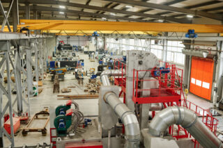 Wide-angle view of large manufacturing plant filled with various heavy machinery and equipment. Several workstations are visible with an overhead crane system installed